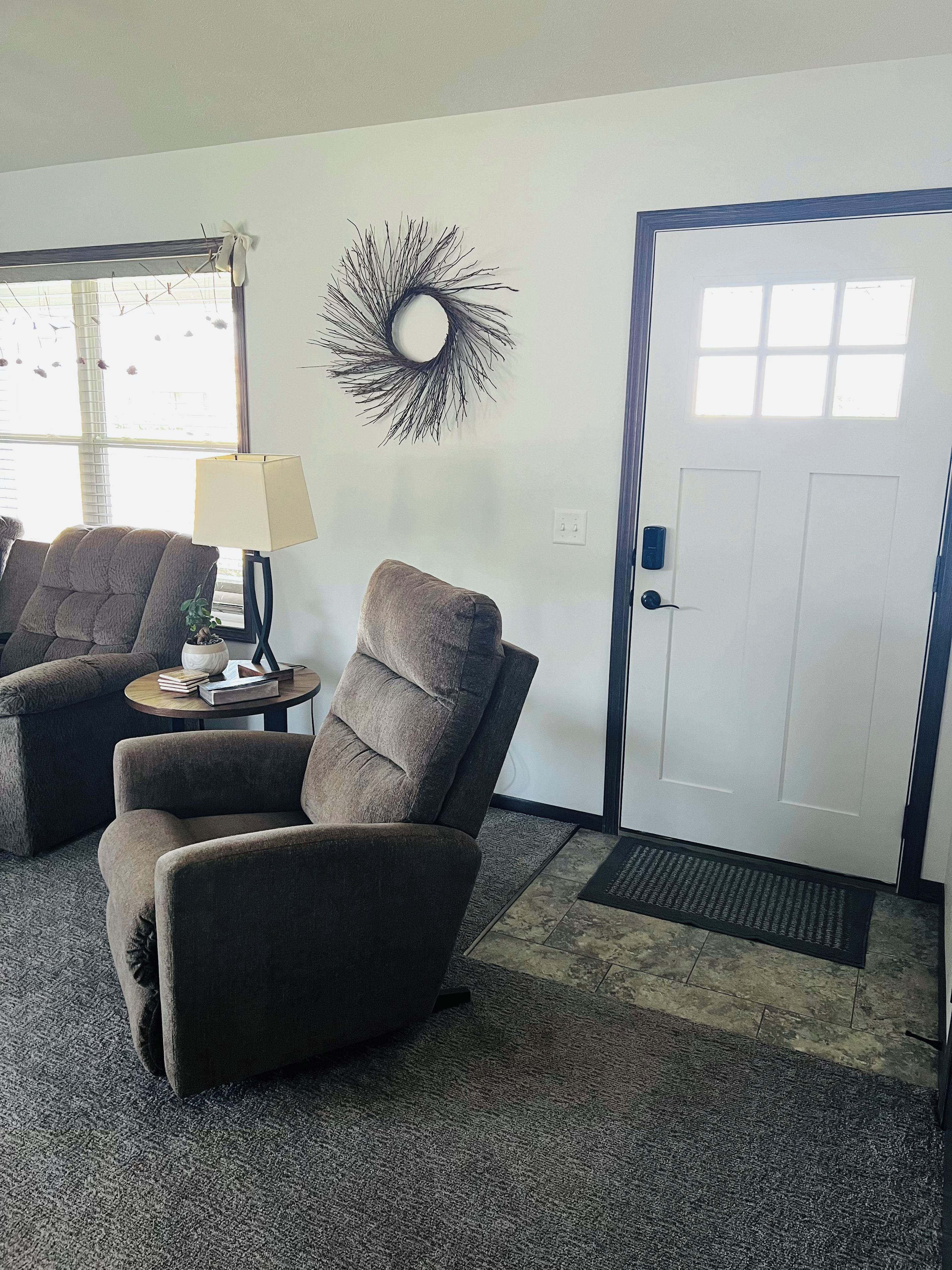 A living room with two armchairs, a small table with a lamp, a decorative wall piece, and a door with a doormat.