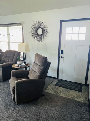 A living room with two armchairs, a small table with a lamp, a decorative wall piece, and a door with a doormat. -  HARVEST COTTAGE Cozy home for family memories in a small town.. (Hesston)