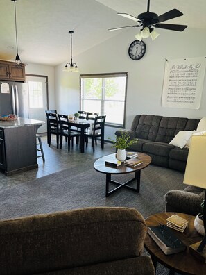 A living room with a sofa, a coffee table with books, a dining area with chairs and a table, a kitchen with cabinets and a refrigerator, and a ceiling fan. -  HARVEST COTTAGE Cozy home for family memories in a small town.. (Hesston)