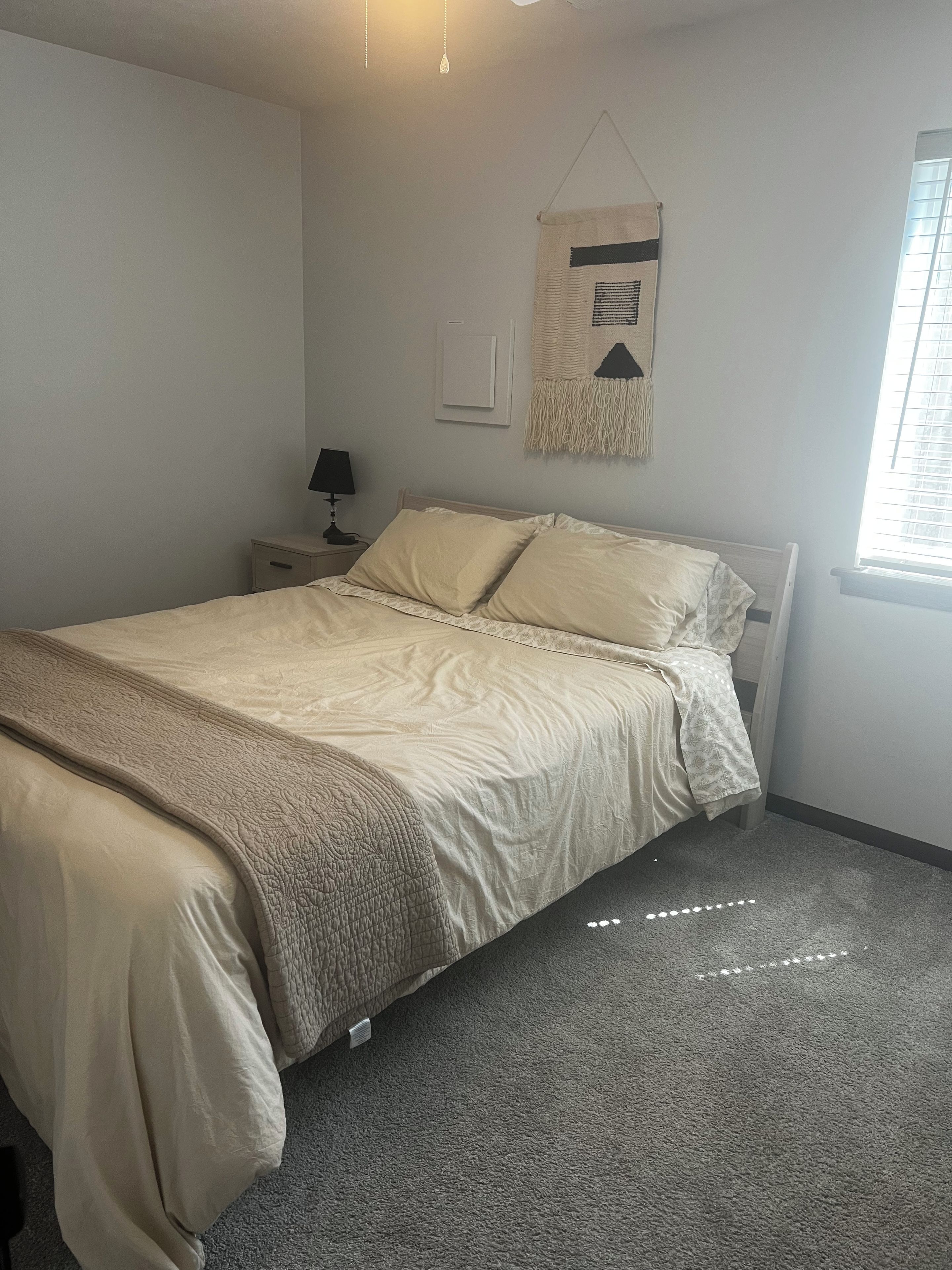 A bedroom with a bed, a nightstand, a lamp, a wall hanging, and a window.