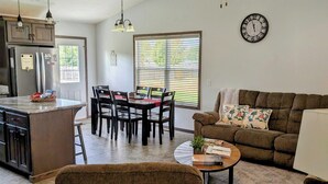 A spacious living room with a sofa, coffee table, and a dining area with chairs and a table. A ceiling fan and a clock are visible on the walls. -  HARVEST COTTAGE Cozy home for family memories in a small town.. (Hesston)