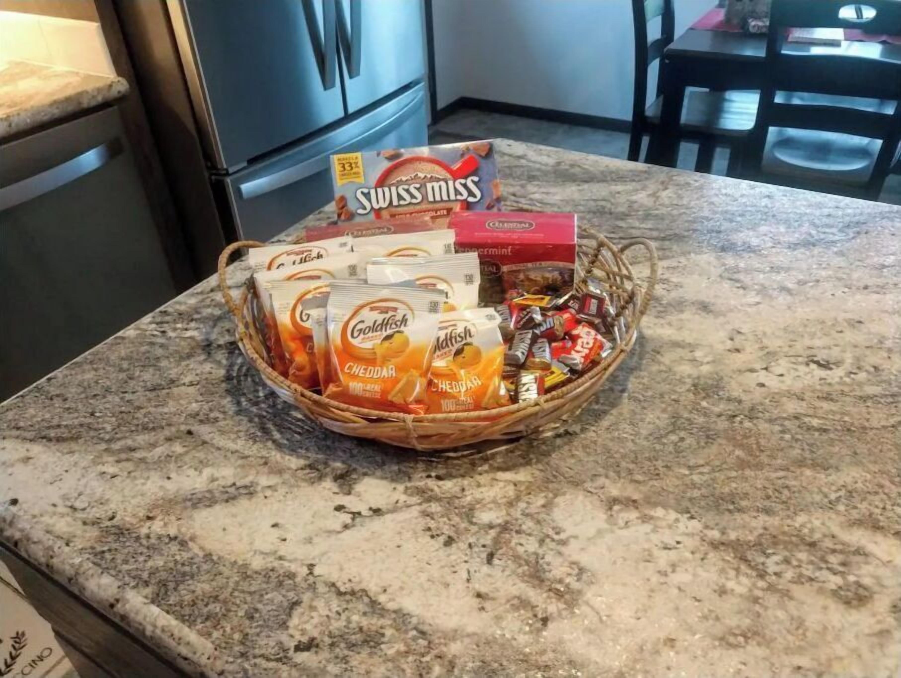 A kitchen counter with a basket containing various food items including Swiss Miss chocolate powder, Goldfish crackers, and other snacks.