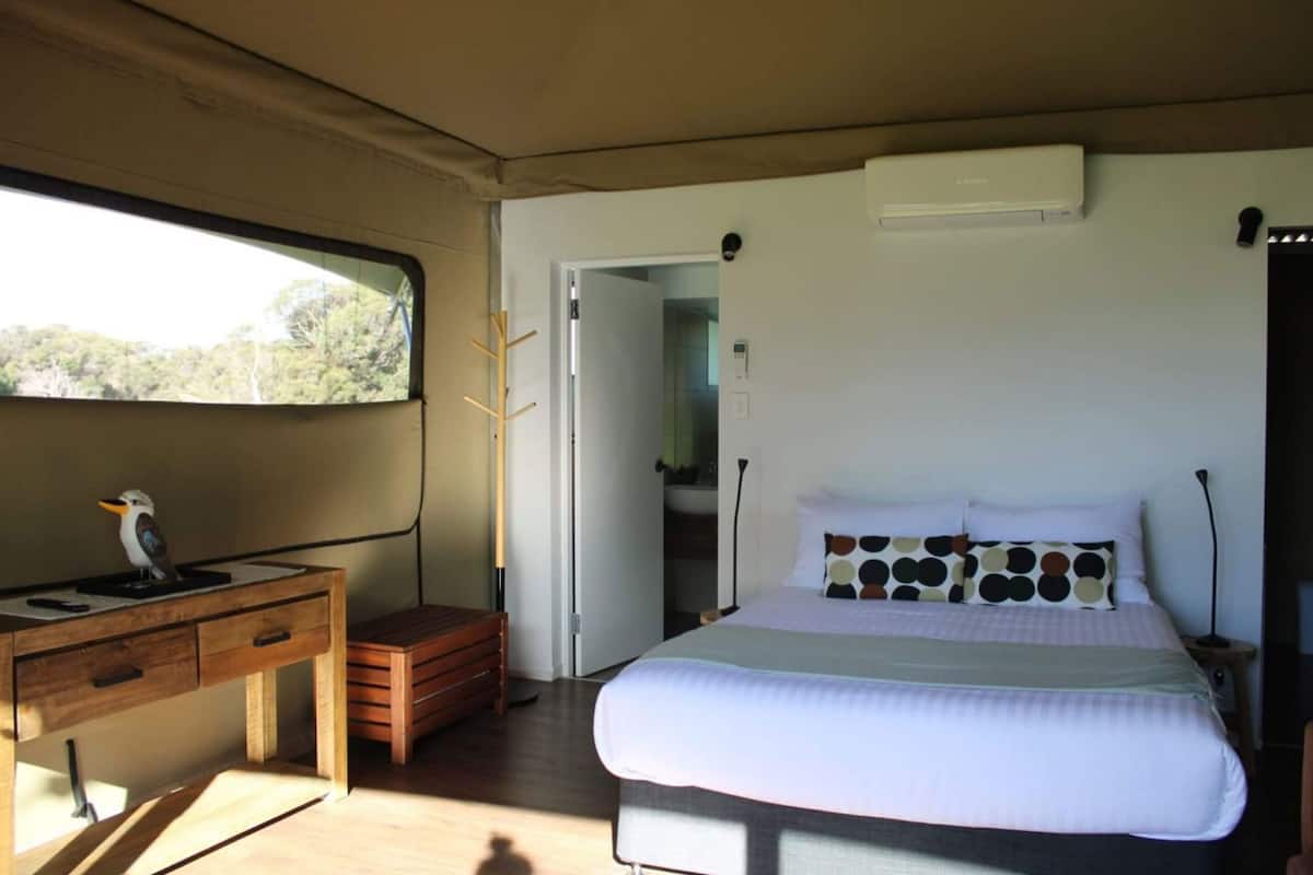 A bedroom with a bed, a desk with a bird, a window, and an air conditioning unit.