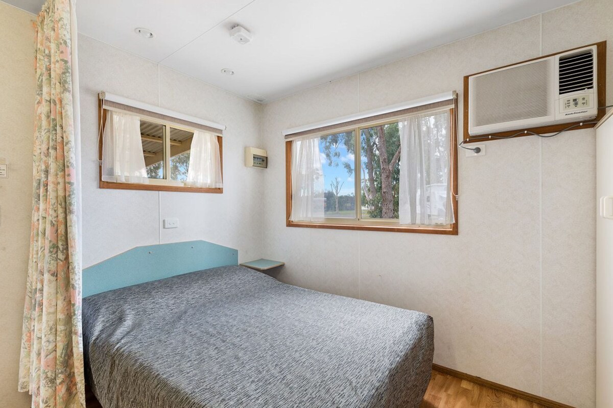 Un dormitorio con una cama, dos ventanas, un aire acondicionado de pared y una mesita de noche pequeña.