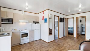 Cocina compacta con electrodomésticos blancos, microondas y estufa. Cuenta con un pequeño comedor con mesa y sillas. El ambiente tiene suelo de madera y un armario con puerta espejada.