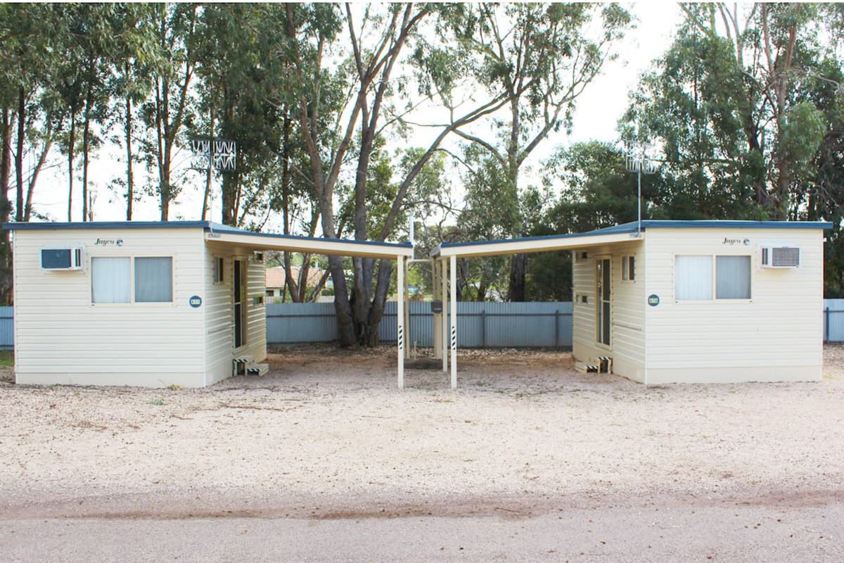 Two small, single-story mobile homes with a covered outdoor area, surrounded by trees.