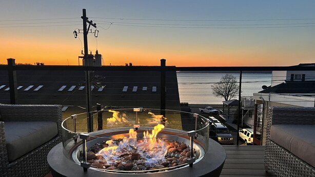 Ein Lagerfeuer auf einer Holzterrasse mit Blick auf Wasser und Gebäude im Hintergrund.