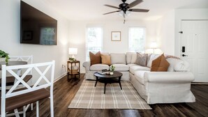 A living room with a white sofa, a black coffee table, and a ceiling fan. There is a white door and a window with blinds. - TigerTown Retreat - Comfort Minutes from Campus (Central)