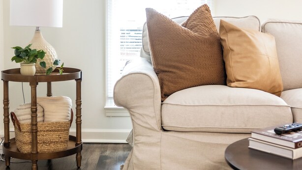 A living room with a beige sofa, a round wooden side table with a lamp, a small wicker chair, and a patterned rug. - TigerTown Retreat - Comfort Minutes from Campus (Central)