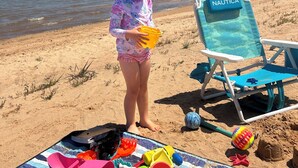 Eine Strandansicht mit einem Kind, das ein gelbes Objekt hält, einem blauen Liegestuhl mit der Aufschrift 'NAUTICA' und verschiedenen Strandspielzeugen auf einer Decke.