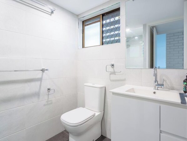 Un baño moderno con azulejos blancos, inodoro, lavamanos y ventana con reja de metal.