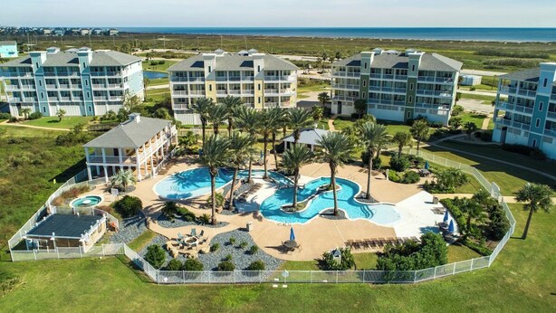 A resort with multiple buildings, a central pool area with palm trees, and a view of the ocean. - "Above the Waves" - BEACH FRONT (Galveston)