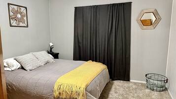 Un dormitorio con cama, mesita de noche, lámpara, ventana con cortinas, decoración en la pared y piso alfombrado.