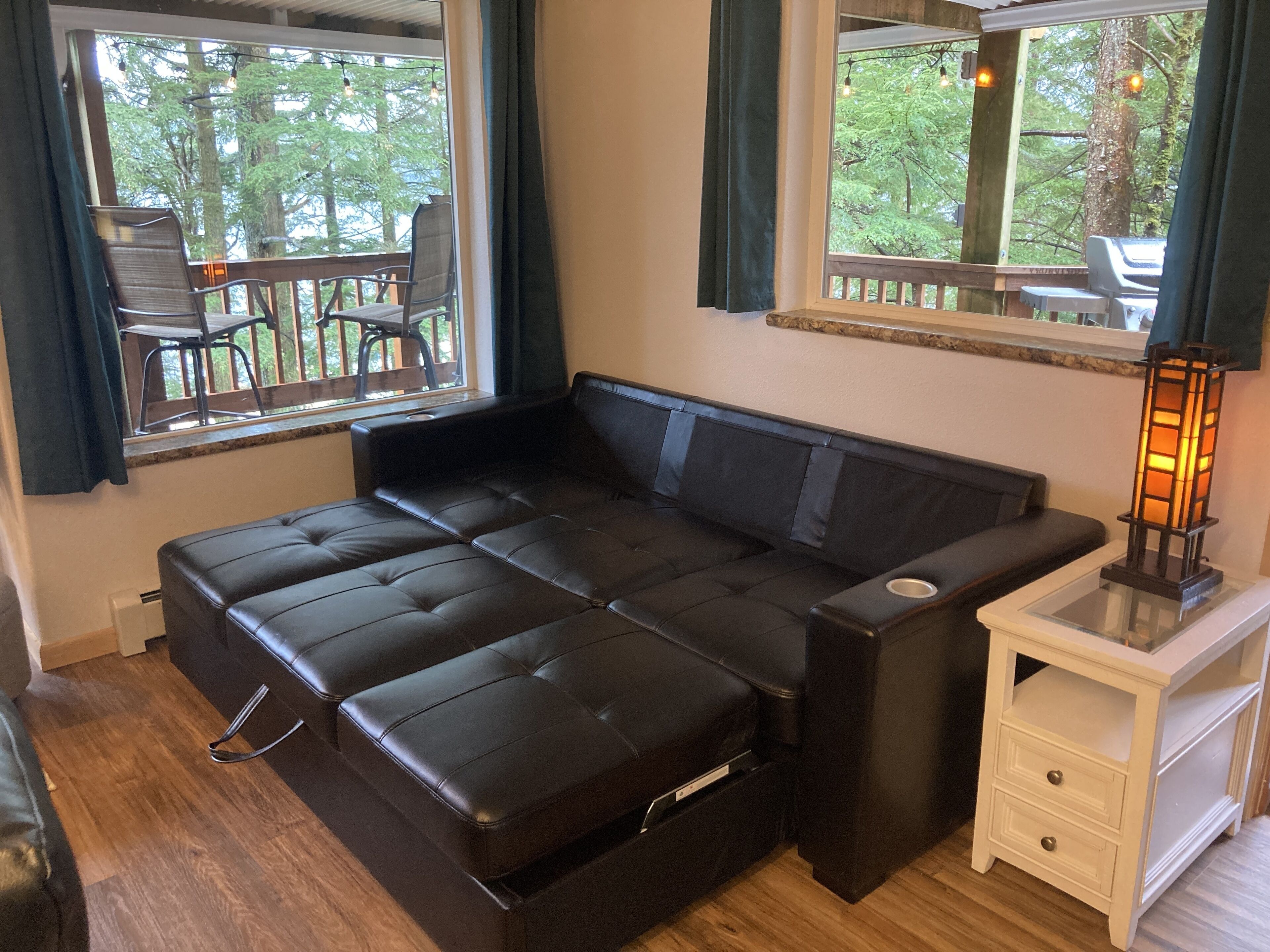 A modern room with a black leather sofa, a small white nightstand, and a window overlooking a deck with outdoor furniture.