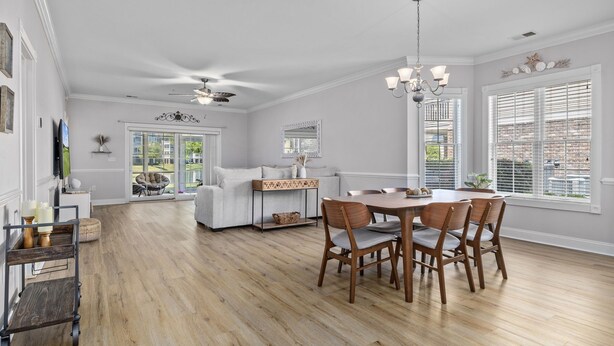 A spacious dining area with a wooden table and chairs, a chandelier, and a large window with blinds. - Cozy Condo//Pool/Workspace/Dogs Stay for Free! (Myrtle Beach)