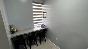 Una habitación pequeña y estrecha con una ventana con persianas, una mesa blanca y tres sillas negras.