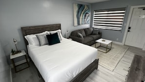 A bedroom with a large bed, a grey sofa, a small table, and a painting on the wall. - Cozy Beachside Modern Boutique Hotel Suite with Kitchen - by Oceano Suites (Daytona Beach)