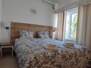 A bedroom with a wooden headboard, a bed with floral bedding, two bedside lamps, a nightstand, and a window with curtains. - Villa 81, 'cozy', in a fantastic location on Oura beach, Albufeira (Albufeira)