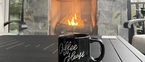 A black coffee mug with "Coffee please" on a coaster at a table with a stone fireplace in the background.