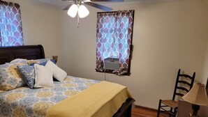 Une chambre à coucher avec un lit, un ventilateur de plafond, une fenêtre avec des rideaux, une table avec une lampe et une chaise.
