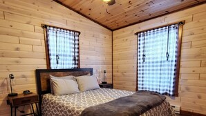 Um quarto de cabana de madeira aconchegante, com uma cama, duas mesas de cabeceira, um ventilador de teto e janelas amplas com cortinas xadrez.