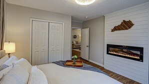 A bedroom with a bed, a bedside table with a lamp, a dresser, and a fireplace.