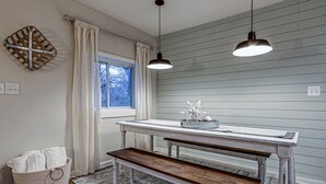 A room with a white table, a bench, a window with white curtains, and two pendant lights.