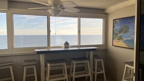 Een kamer met een groot raam met uitzicht op de oceaan, een plafondventilator en een klein huismodel op de vensterbank.