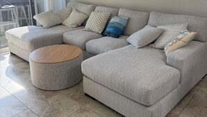 Een moderne woonkamer met een hoekbank, een ronde salontafel en een groot raam met gordijnen.