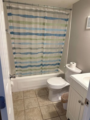 Une salle de bain avec un rideau de douche à rayures, des toilettes blanches, une baignoire, un lavabo blanc et une poubelle.