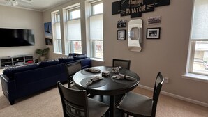 Un salon moderne avec un canapé d’angle bleu foncé, une table de salle à manger ronde entourée de quatre chaises, un ventilateur de plafond et un téléviseur à écran plat fixé au mur.