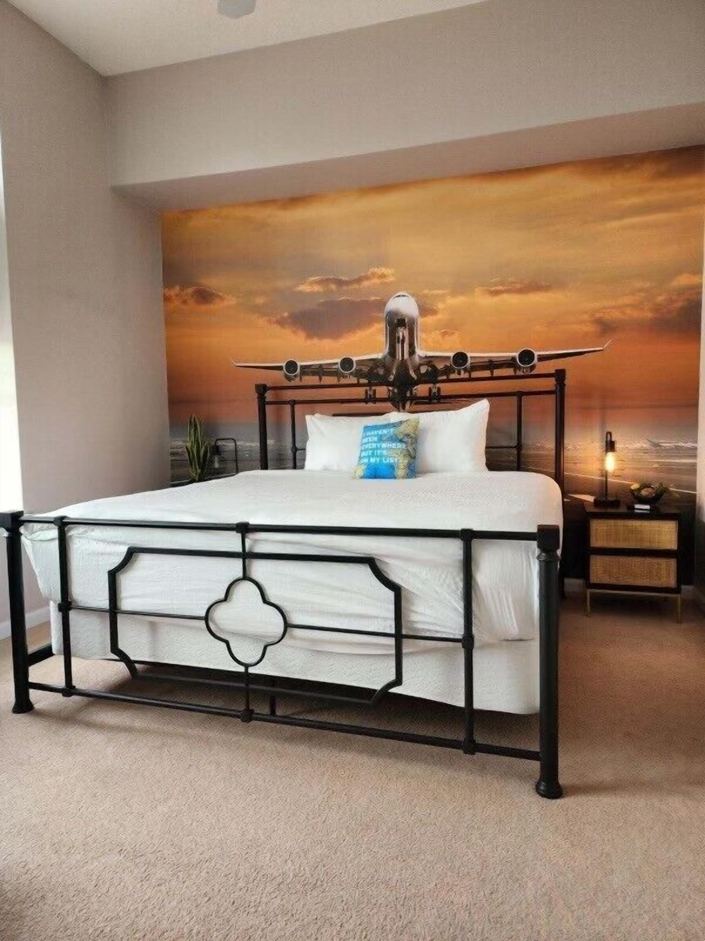 Ein Schlafzimmer mit einem großen Bett, einem Nachttisch, einer Lampe und einem Wandbild, das einen Sonnenuntergang mit einem Flugzeug zeigt.
