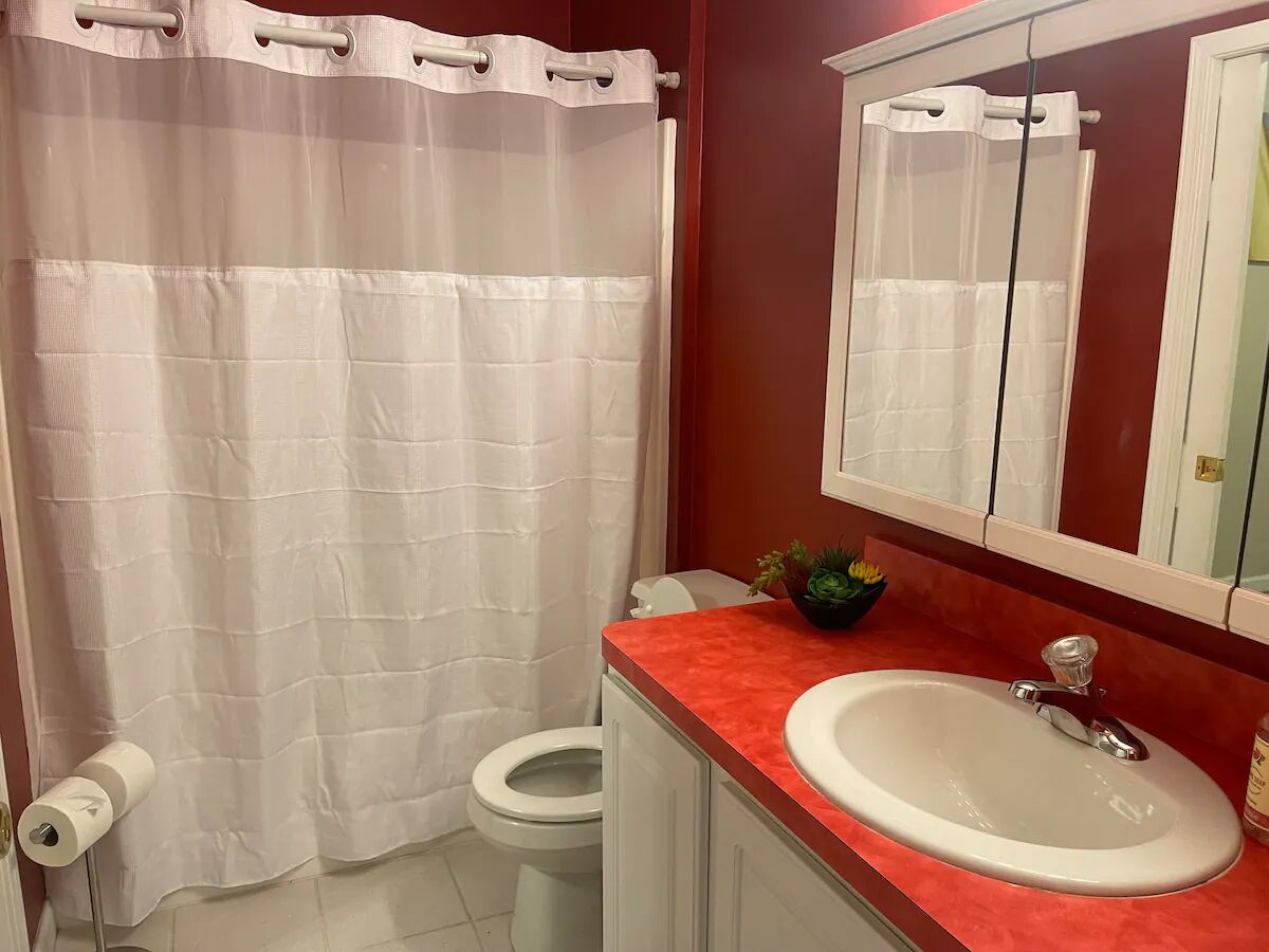 Ein Badezimmer mit Toilette, einem weißen Waschbecken mit roter Ablage, einer weißen Duschvorhang und einem Spiegel über dem Waschbecken.
