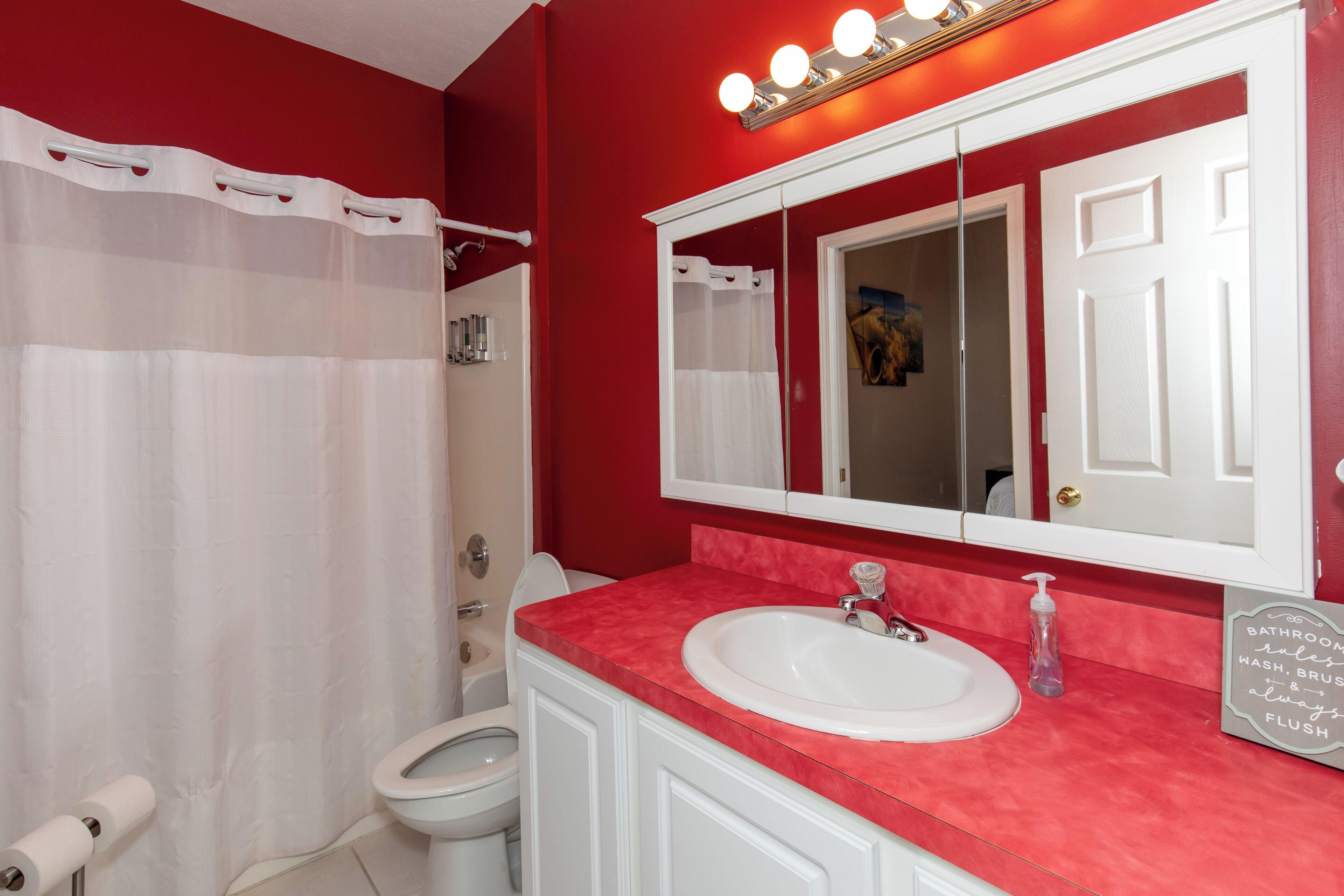 Un baño con pared roja, sanitarios blancos, un espejo grande y un cartel que dice 'bathroom rules'.