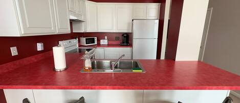 A compact kitchen with white cabinets, a red countertop, a sink, a microwave, a toaster, and a refrigerator. There are two chairs at the island.