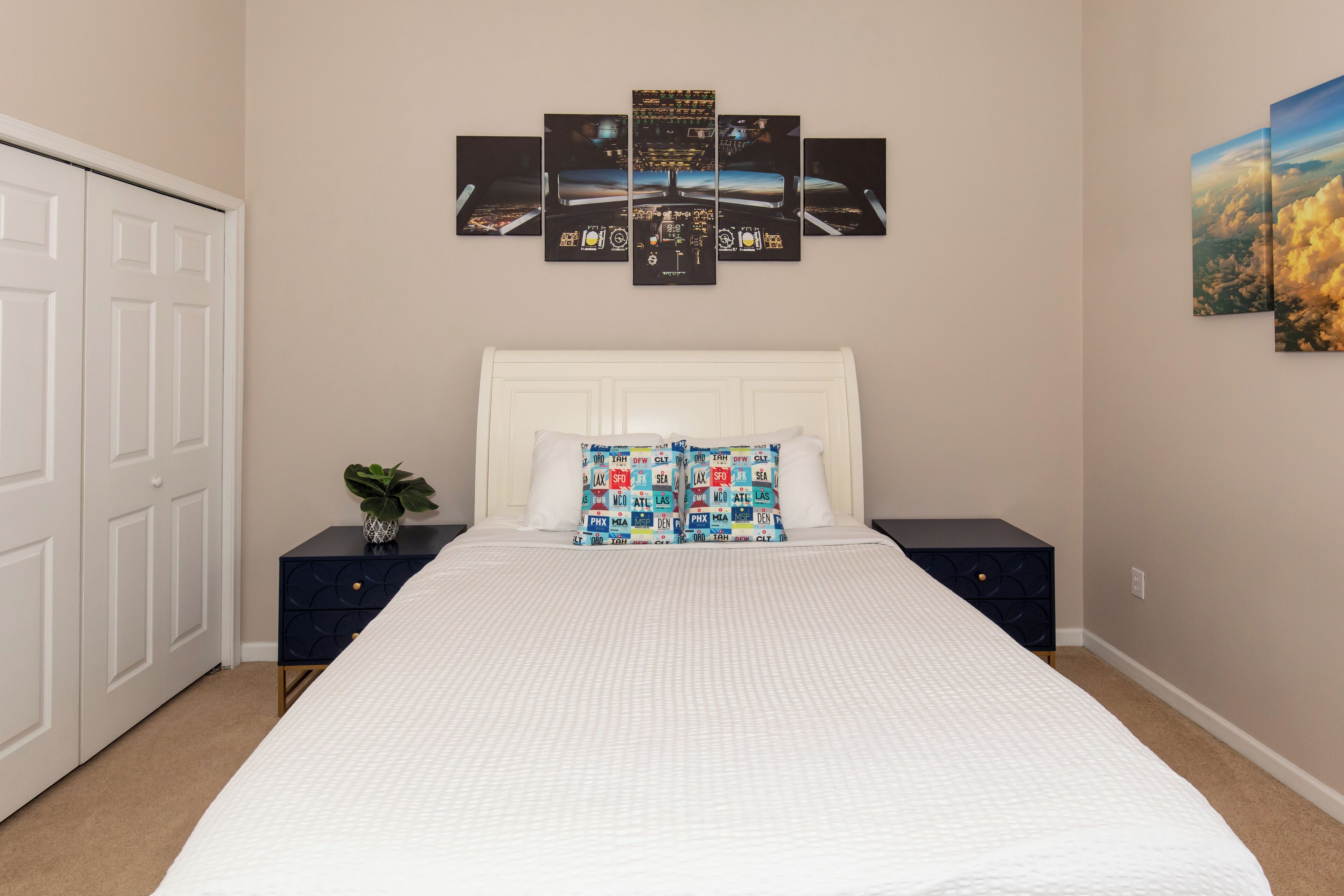 A bedroom with a large bed, a white headboard, a multi-panel artwork above the bed, a small plant on a nightstand, and a closet with sliding doors.