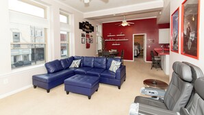 Un salon avec un canapé d’angle bleu, un fauteuil inclinable gris et un ventilateur de plafond.