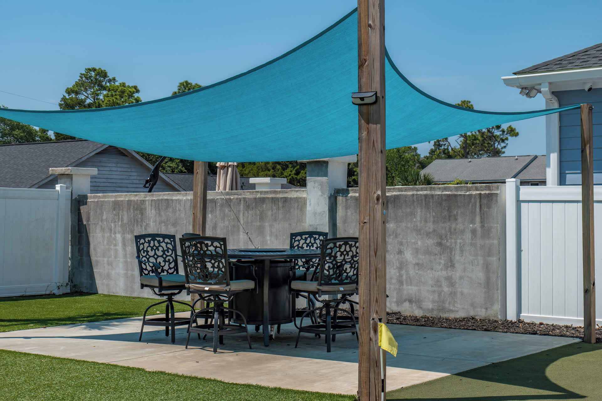 An outdoor patio with a table and chairs, a blue shade sail, and a white fence.