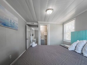 A bedroom with a bed, a window, a door leading to a bathroom, and a painting on the wall. - The Seven Oh Five | Coastal Duplex (North Myrtle Beach)