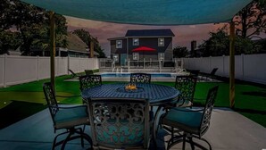 A covered outdoor patio with a round table and chairs, a pool, and a house in the background.