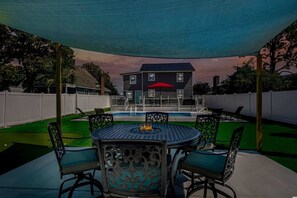Une terrasse extérieure couverte avec une table ronde et des chaises, une piscine et une maison en arrière-plan.