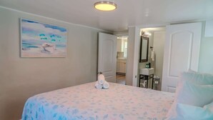 Quarto com cama, pintura de uma praia, banheiro com pia e espelho, e porta que dá para outro cômodo.