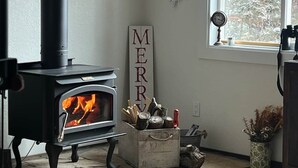 A wood-burning stove with a fire, a 'MERRY' sign, a snowflake decoration, and a window with a view of trees. - HOT TUB ~ 31 Acres ~Bring ATVs/Border Nat'l Forest (Teller County)