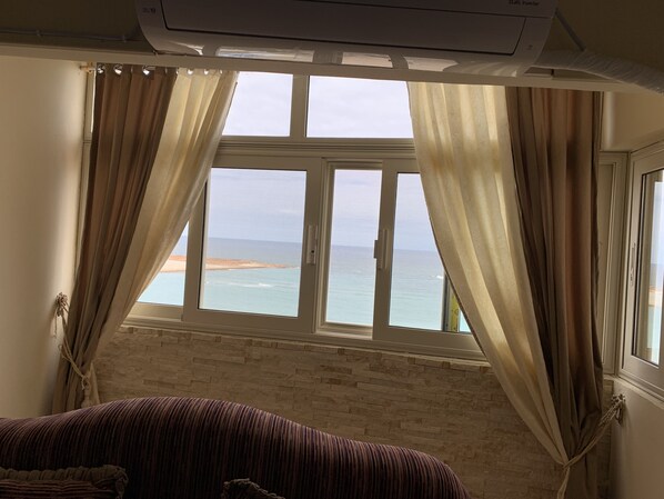 A room with a large window overlooking a beach, a brick wall, and a bed with a striped blanket.