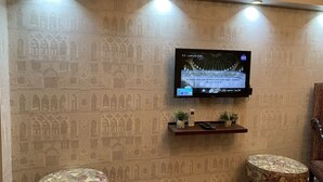 Una habitación con un papel tapiz decorativo, un televisor montado, dos otomanes y un pequeño estante con un control remoto.
