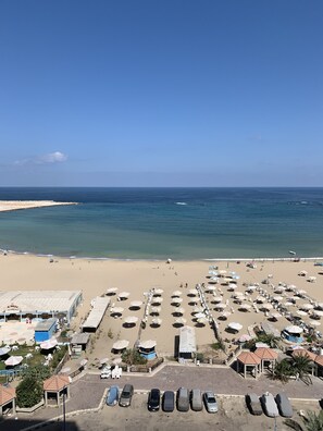 A beach with numerous umbrellas and sun loungers, a parking area, and a view of the ocean. - Apartment with Stunning Mediterranean Seaside View (Alexandria Governorate)
