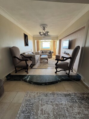 A living room with a sofa set, two armchairs, a coffee table, a television, and a view of the sea through a window. - Apartment with Stunning Mediterranean Seaside View (Alexandria Governorate)
