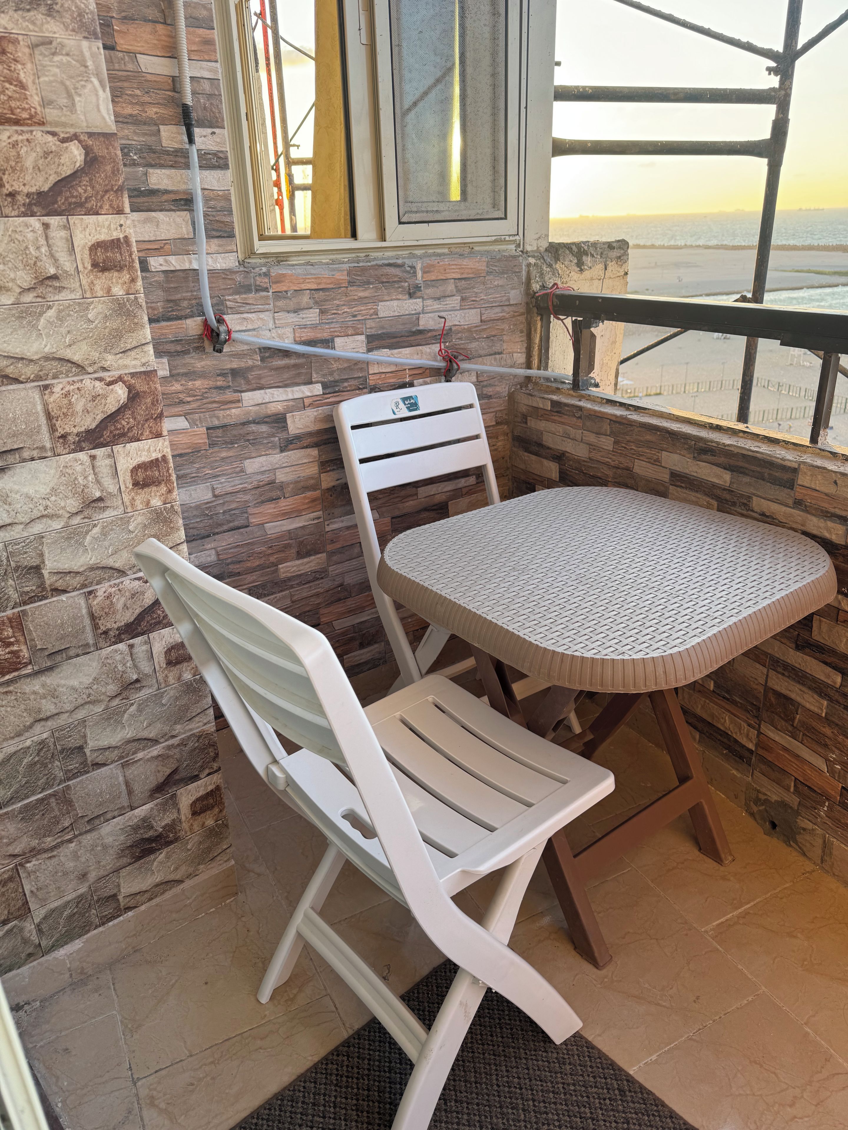 A small outdoor seating area with a table and two chairs, a window with a view of the beach, and a stone wall.