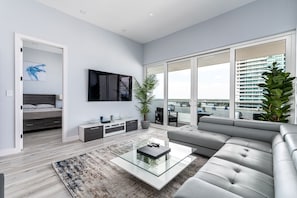 Uma sala de estar moderna com um sofá cinzento, uma mesa de centro de vidro e uma televisão grande na parede.