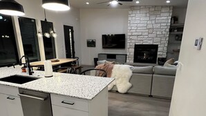 Una cocina moderna con una isla central, un comedor con mesa y sillas, una sala de estar con sofá y chimenea, y un ventilador de techo.